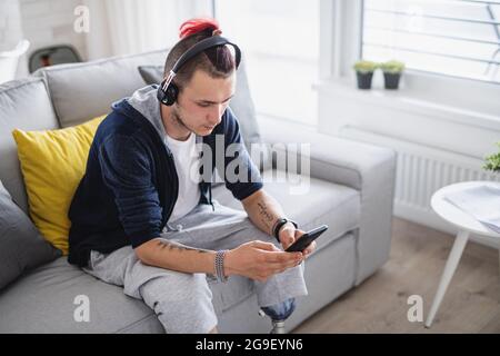 Portrait of disabled young man using smartphone indoors at home, leg prosthetic concept. Stock Photo