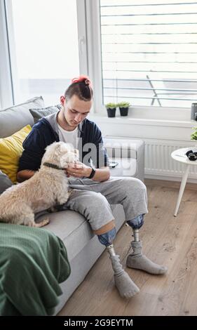 Portrait of disabled young man playing with dog indoors at home, leg prosthetic concept. Stock Photo
