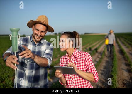 Female farmer using tablet to monitor rainfall. Young male farmer holding up rain gauge pointing explaining. Man using portable hand sprayer on plants Stock Photo