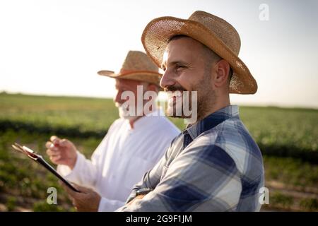 Young man farmer standing in field with arms crossed smiling. Elderly scientist wearing white coat holding clipboard taking notes. Stock Photo