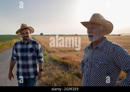 Young male farmer wearing plaid shirt and hat looking at camera. Elderly male farmer standing in field with hands on hips looking to side. Stock Photo