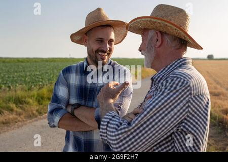 Young and elderly farmers standing on road next to field with crossed arms. Two men wearing plaid shirts and straw hats talking laughing. Stock Photo