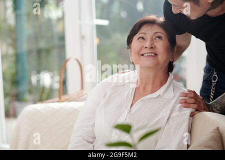 Asian matue woman sitting on sofa smiling and talking to her elder son at home Stock Photo