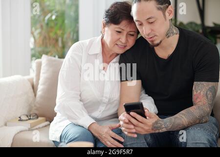 Young man showing something on his mobile phone to his mature mother while they sitting on sofa at home Stock Photo