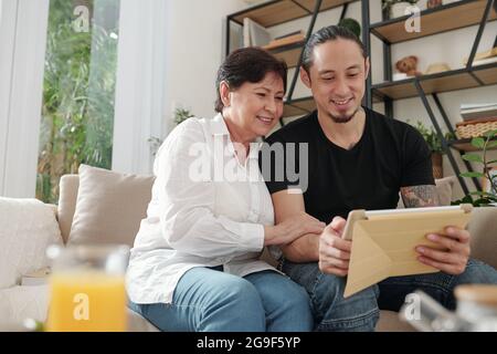 Smiling elder son watching something on digital tablet together with his mother on the sofa in the room Stock Photo