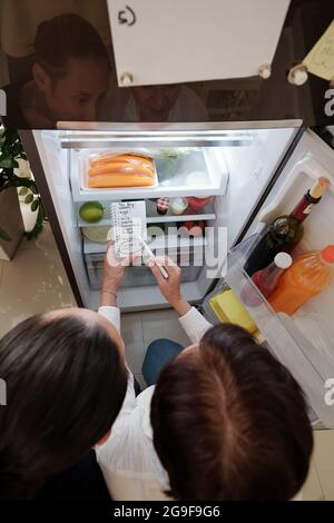 Rear view of mother and son standing in front of the opened fridge and making shopping list together Stock Photo