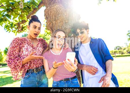 Three diverse students making surprised faces spreading mouth and eyes pointing and looking smartphone spending time in nature of city park. power of Stock Photo
