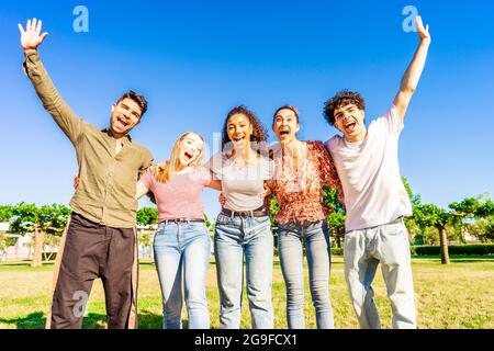 Multiracial group of young friends posing with raised open arms looking at camera embracing to each other in nature of city park. Happy diverse people Stock Photo