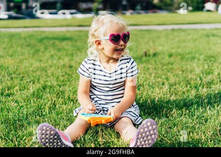 Adorable lively little blonde girl in dress and sunglasses sits on a grass and cheerful smiling. Stock Photo