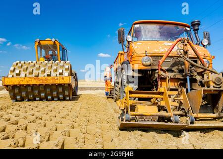 Road roller with spikes and truck with mounted plate vibration compactor are compacting, leveling sand for road foundation at building site. Stock Photo