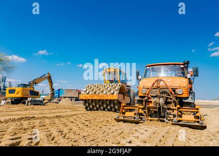 Road roller with spikes and truck with mounted plate vibration compactor are compacting, leveling sand for road foundation at building site. Stock Photo