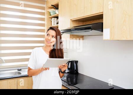 Smiling pretty young woman standing in kitchen and using smart home app on her digital tablet when setting temperature Stock Photo