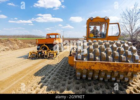 Road roller with spikes and truck with mounted plate vibration compactor are compacting, leveling sand for road foundation at building site. Stock Photo