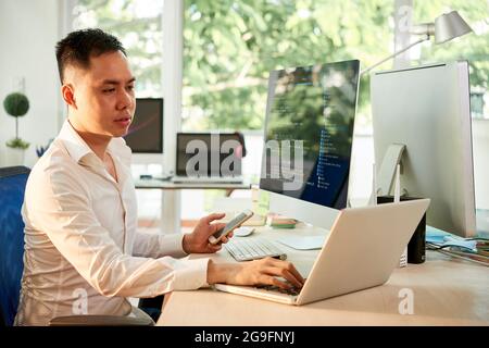 Young software engineer with smartphone in hand working on laptop when checking programming code Stock Photo