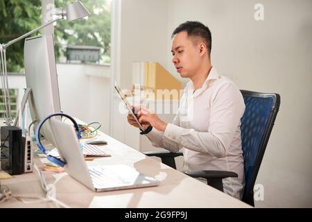 Young software engineer sitting at office desk and testing new application he was working on on tablet computer Stock Photo