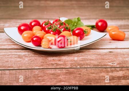 Mixed Yellow And Red Cherry Tomatoes Lie With Leaves Of Basil On A White Plate And Rough Brown Wooden Table. Fehraltorf, Switzerland Stock Photo