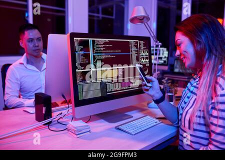 Smiling female software developer sitting at office desk and texting friends instead of coding Stock Photo
