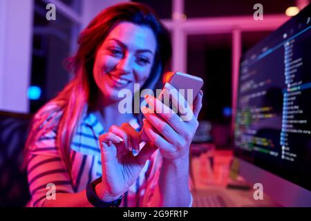 Cheerful female coder working late at night in office with neon light and texting friends, focus on hands Stock Photo