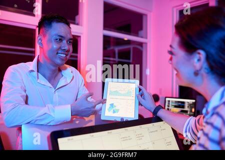Positive entrepreneurs discussing diagram on tablet computer showing good sales when working in neon light Stock Photo