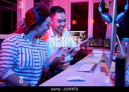 Coders having short break and discussing funny memes and videos on digital tablet, pink neon light Stock Photo
