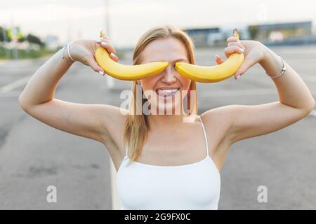 Frivolously and mischievous young woman with braces on teeth holding bananas on her face has fun outdoors. Emotions Stock Photo
