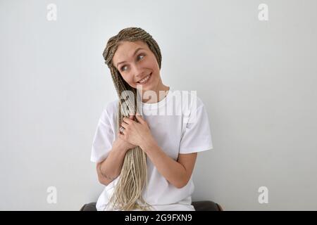 Positive female wearing white t shirt sitting near wall and touching braids while looking away Stock Photo
