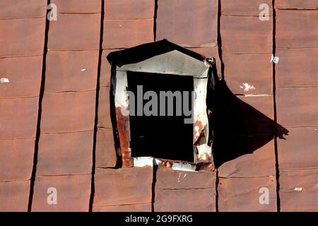 Twisted damaged broken old roof window frame without glass on top of abandoned suburban family house roof covered with rusted cracked metal roof tiles Stock Photo