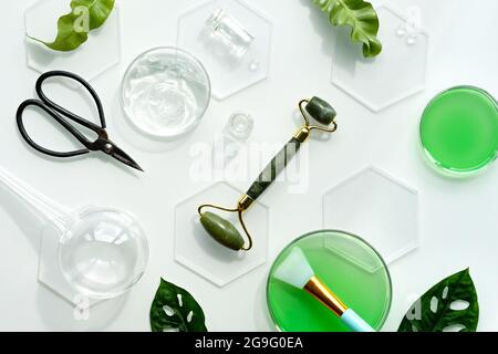Self made moisturizer, green jade face roller and scissors with pieces of ice. Exotic monstera leaves and water drops on white background. Facial mass Stock Photo