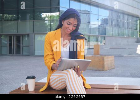 Young indian businesswoman wearing yellow suit jacket working with a tablet sitting outdoor. Stock Photo
