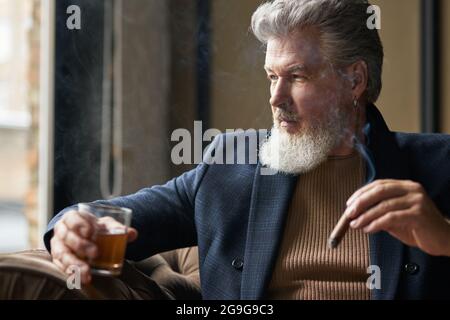 Portrait of thoughtful bearded mature businessman holding cigar and a glass of whisky while relaxing in loft interior Stock Photo