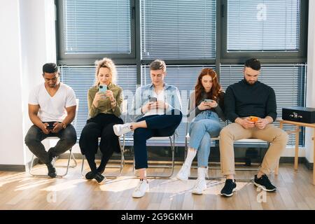 Front view of group five young diverse ethnicity candidates using mobile phones while waiting job interview in modern office lobby. Stock Photo
