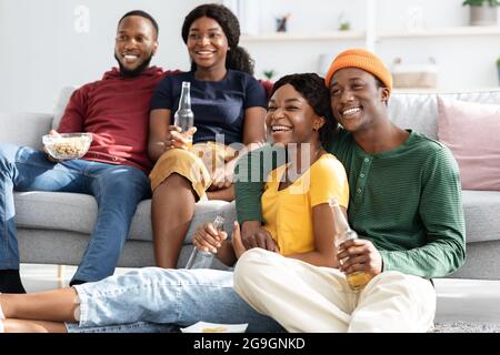 Joyful african american friends sitting on floor, watching movie and drinking beer at home, copy space. Happy group of young black men and women two c Stock Photo