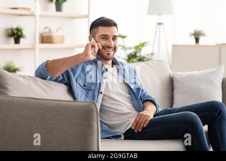 Happy cheerful young man talking on the phone at home, smiling jewish male making or answering call by cellphone sitting on sofa, handsome guy having Stock Photo