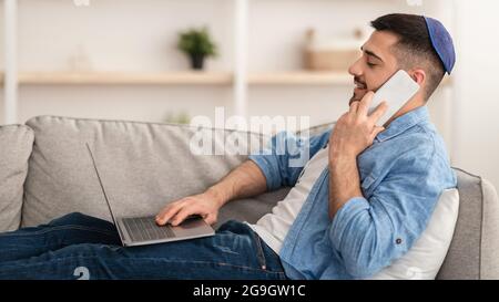 Home Office Concept. Side view profile portrait of smiling millennial man talking on smart phone using pc at workplace, sitting on sofa. Young male em Stock Photo