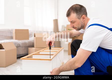 Professional Furniture Assembler Man Fixing And Assembling Wooden Shelf After Relocation Sitting On Floor Indoors. Side View Of Carpenter Working Usin Stock Photo