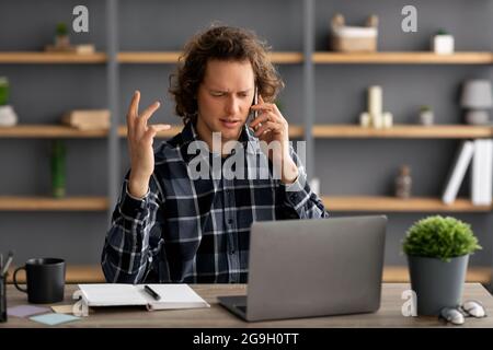 Displeased Businessman Talking On Cellphone Sitting At Laptop Having Problem At Workplace In Modern Office. Unpleasant Phone Conversation, Negative Ca Stock Photo