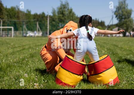 Child in inflatable boots. The kindergartner plays at the party. Boots for funny walking. Uncomfortable shoes on the child. Preschooler game on the gr Stock Photo