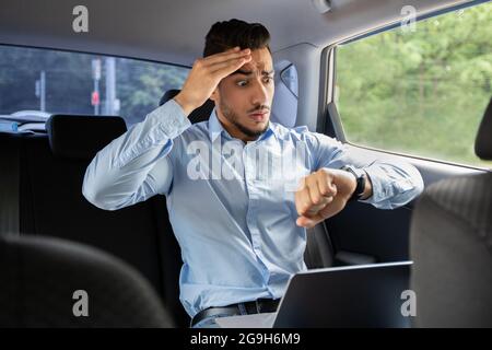 Worried arab guy businessman looking at watch and touching his head while sitting in car bak seat with laptop, shocked middle-eastern entrepreneur get Stock Photo