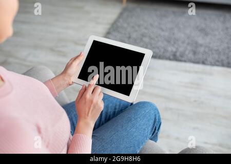 Young lady using tablet computer with empty screen for your website or app advertisement, sitting on sofa in living room interior. Mockup. Internet an Stock Photo