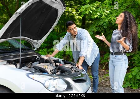 Upset middle-eastern couple having broken car while countryside trip, puzzled arab guy checking engine, emotional brunette lady looking up, sad mixed Stock Photo