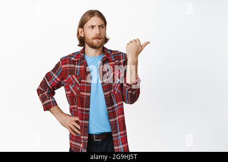 Confused blond man pointing right with frowned face, see smth strange, point at advertisement with puzzled hesitant expresson, standing against white Stock Photo