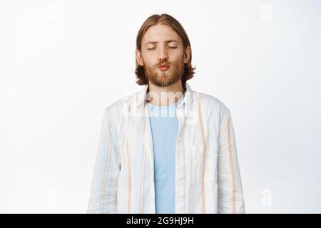 Cute blond bearded guy pucker lips with eyes closed, waiting for kiss, kissing, showing romantic affection, standing over white background Stock Photo