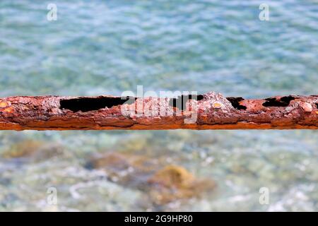 Remains of a rusted handrails on an abandoned waterfront on a blurred background of azure sea waves. Metal corrosion close-up. Stock Photo