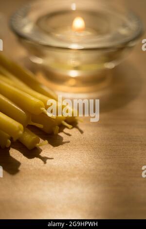 Heap of new long candles on the table. One low candle burns in a glass round candlestick in blurred background. Stock Photo