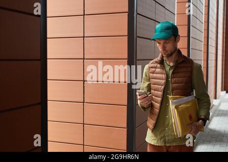 Waist up portrait of delivery man using smartphone while standing in urban setting, copy space Stock Photo