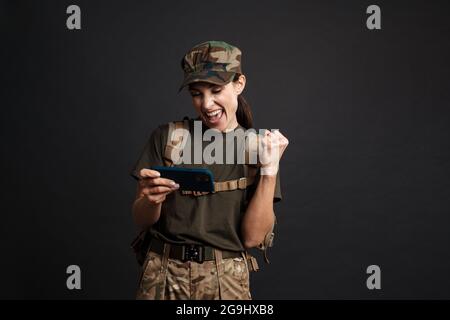 Excited soldier woman making winner gesture while using mobile phone isolated over black background Stock Photo