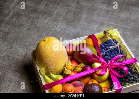 Variety of ripe fruits and berries are beautifully stacked in wooden box tied with pink ribbon. Dark background. Food delivery, gift fruit set. Stock Photo