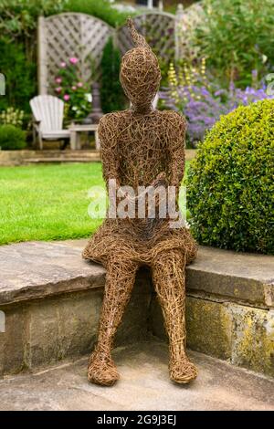 Willow sculpture art of girl (ornamental feature) in beautiful landscaped garden (flowering plants, box ball, lawn, seat) - Yorkshire, England UK. Stock Photo