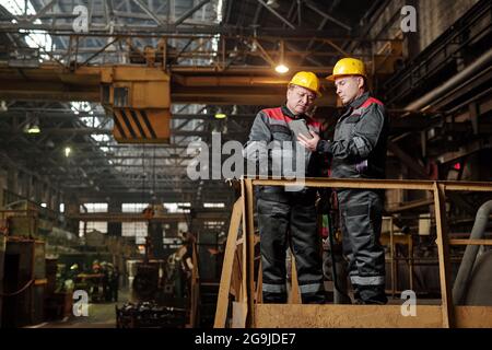 Two manual workers in workwear using digital tablet and discussing work while standing in the plant Stock Photo