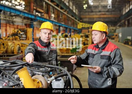 Two workers in helmets using tablet pc and discussing the work of machine equipment in the plant Stock Photo